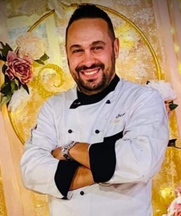Nino Cordova Chef in giacca bianca da cuoco, sorridente a braccia conserte, davanti a un allestimento con fiori e decorazioni dorate