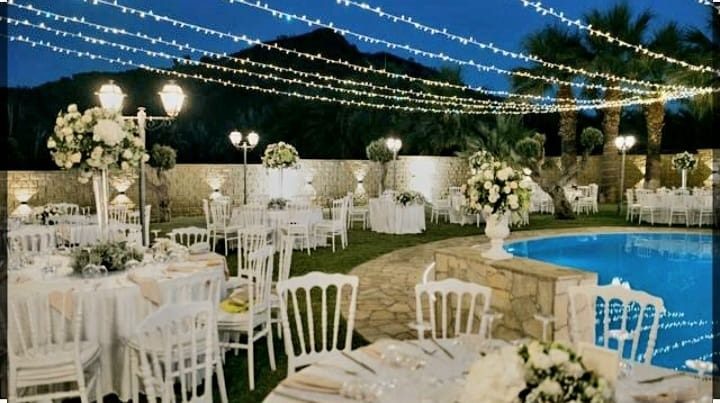 La piscina de L'Antico Podere di sera, allestita per un ricevimento: tavoli con tovaglie bianche e centrotavola di fiori, sedie bianche e luci sospese sopra il prato